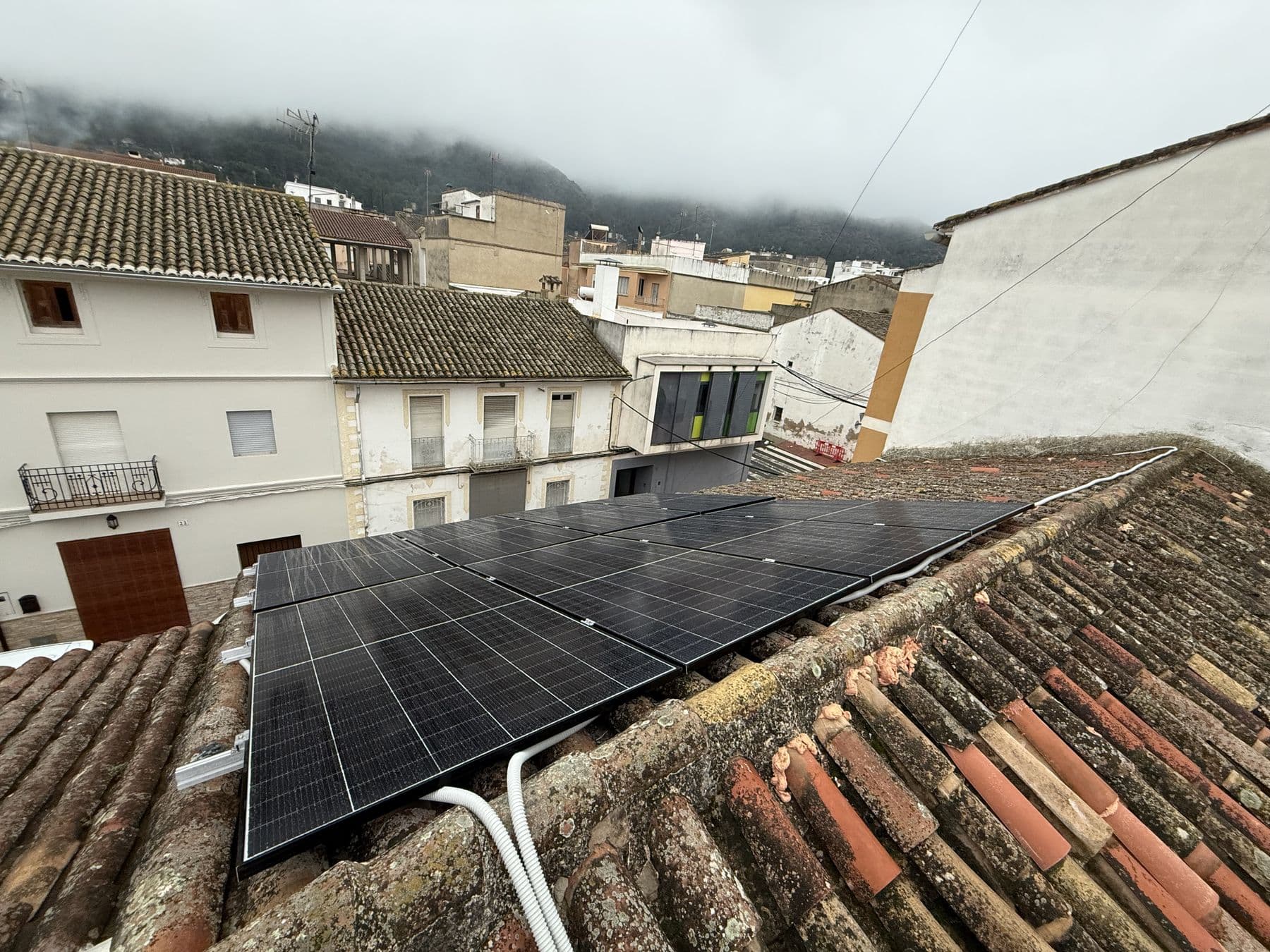 Placas solares coplanares sobre teja árabe en un casco antiguo, con la montaña entre niebla al fondo